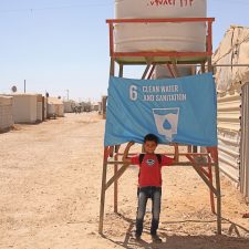 Et barn i Zaâatari flygtningelejren i Jordan står foran et flag, der repræsenterer mål 6: Rent Vand og Sanitet. Foto: Unicef Jordan/Badran